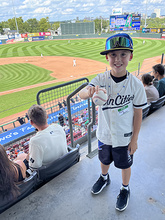 A young child wearing a baseball cap and Twins jersey holds out a baseball while standing in the stadium with the baseball field behind him.