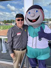 A man in a black baseball cap and sunglasses stands with a costumed character with a baseball head. Blue sky and a baseball field is reflected in the window behind them.