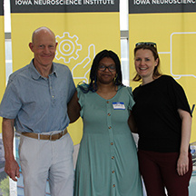 Three people with their arms around each other’s shoulders stand in front of a gold banner with black bar at the top that reads Iowa NeuroScience Institute.