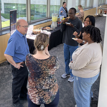 A group of people standing in a circle chatting and eating. In the background is a table with trays of cookies and a drink dispenser full of lemonade.