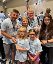 A group of four adults and two children, all wearing matching light blue t-shirts from the Jordan’s Guardian Angels organization, gathers for a photo.