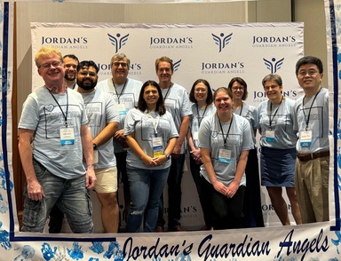 Eleven people in matching light blue t-shirts stand in front of a photo backdrop for the Jordan’s Guardian Angels organization.